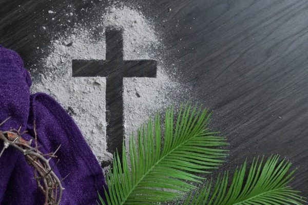 Christian lent background with cross of ashes, palm fronds, purple cloth and partial crown of thorns on a dark wood background with copy space